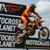 Een motorcrosser op een KTM racemotor vliegt door de lucht tijdens een MXGP-wedstrijd, met banners van Motocross Planet op de achtergrond