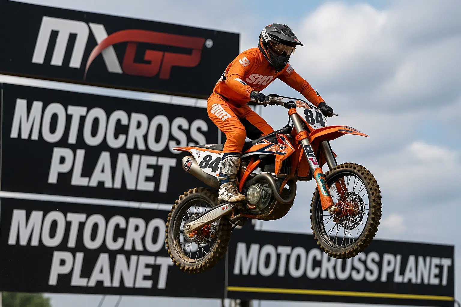 Een motorcrosser op een KTM racemotor vliegt door de lucht tijdens een MXGP-wedstrijd, met banners van Motocross Planet op de achtergrond